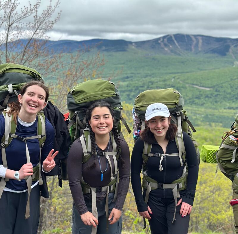 Three young women are posing for a photo while backpacking. They are wearing large backpacks and hiking clothes. The woman on the left is holding up a peace sign. In the background, there is a mountain range and a cloudy sky. The women appear to be enjoying their outdoor adventure.